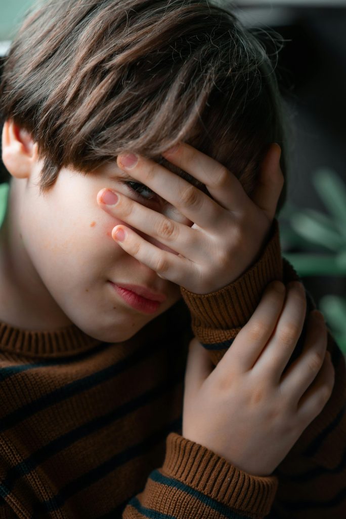 Close-up portrait of a shy young boy covering his face with his hand, indoors.